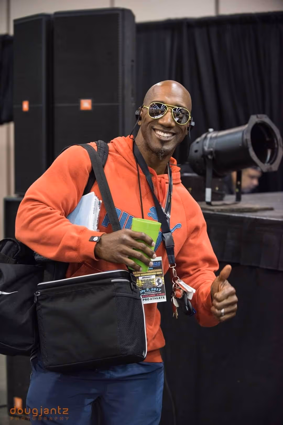 Smiling man in orange hoodie wearing sunglasses gives thumbs up while holding a green cup and keys.