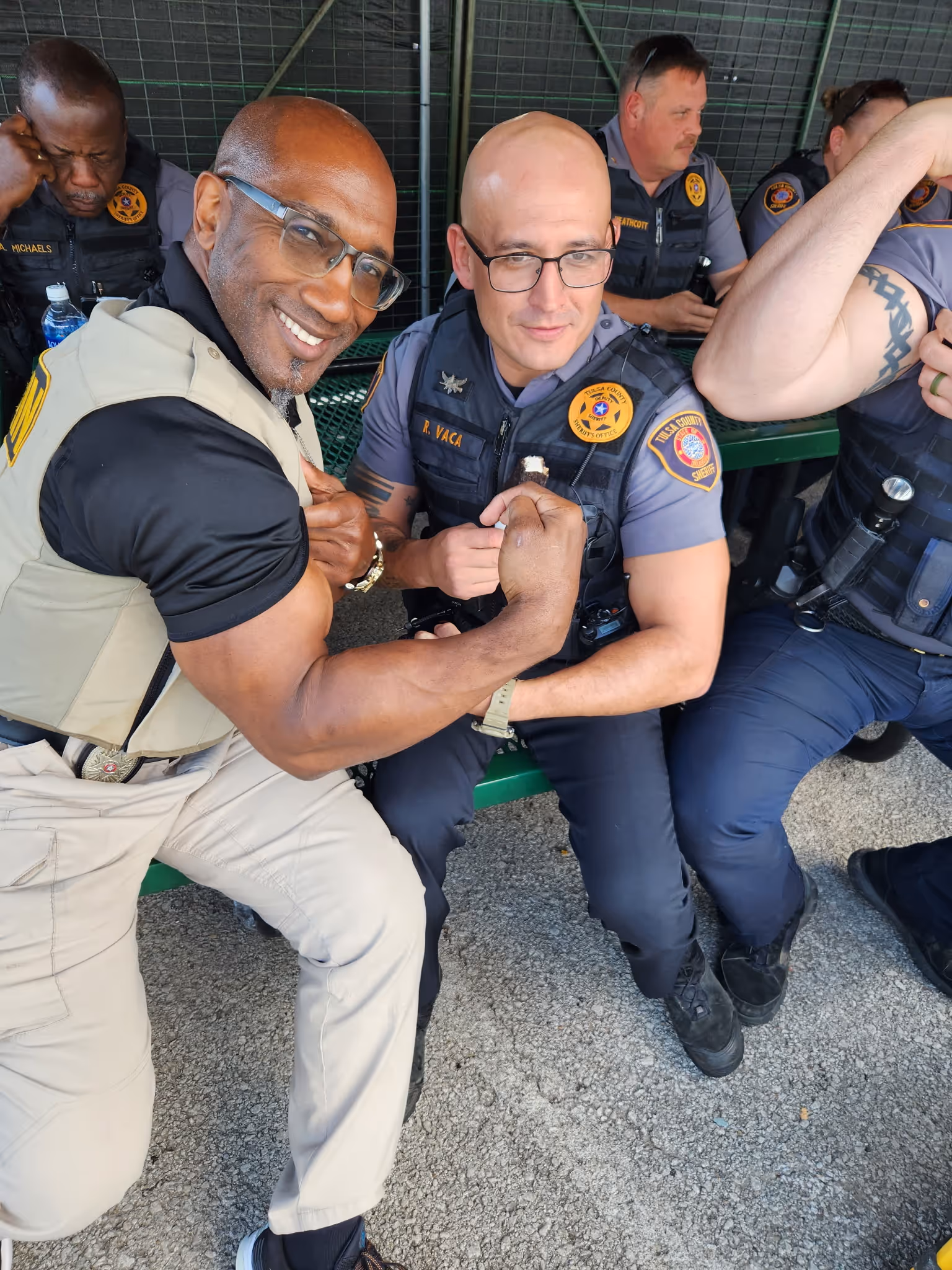 Two men, one in tan uniform vest and the other in Tulsa County sheriff uniform, flex their arm muscles.