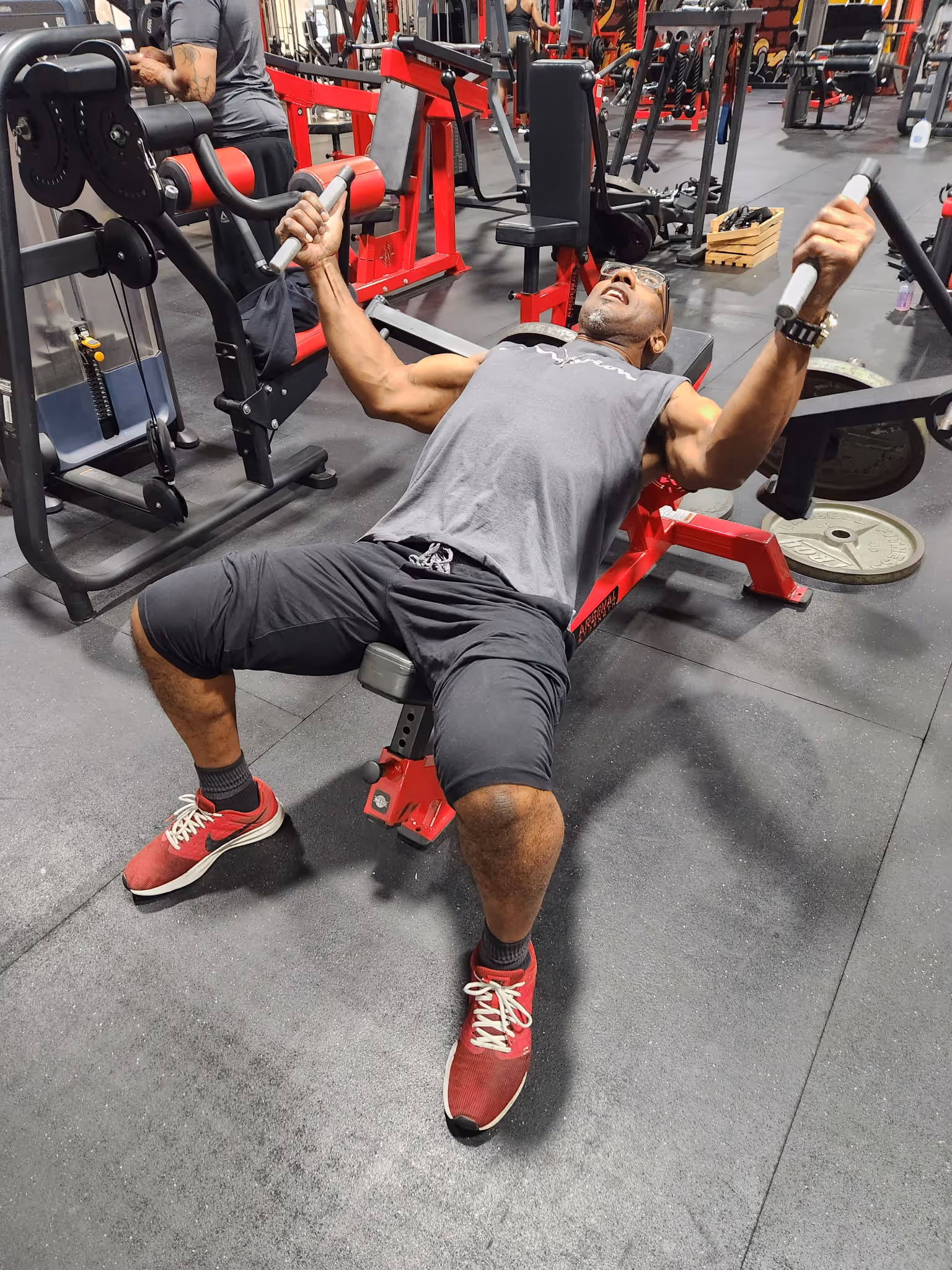 Man lifting weights on an incline chest press machine in a gym wearing red shoes and gray outfit.