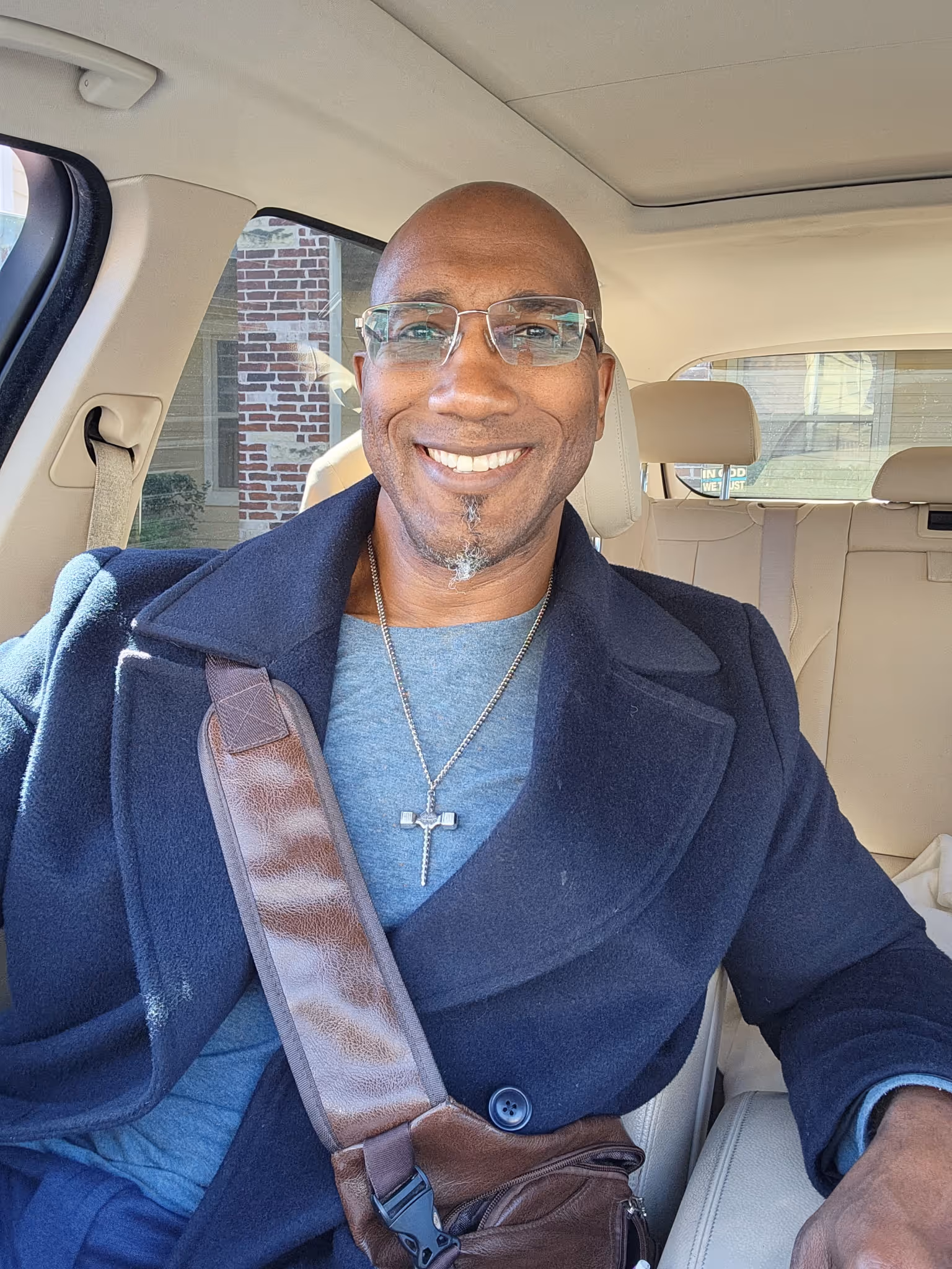 Smiling man with glasses, wearing a coat and cross necklace, seated in a car with beige interior.