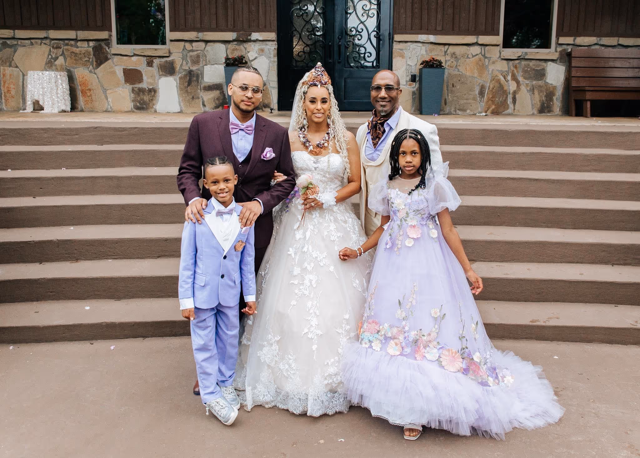 A bride in white lace gown with two children and two men standing on steps outside a stone building.