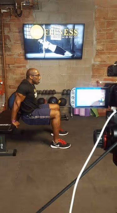 Man doing tricep dips on a bench in a gym with fitness video displayed on a screen behind him.