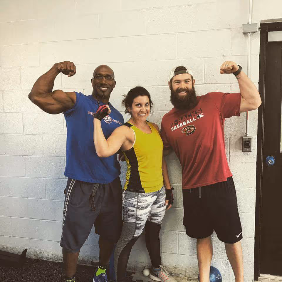 Three fit people smiling and flexing their arm muscles against a white wall in a gym.