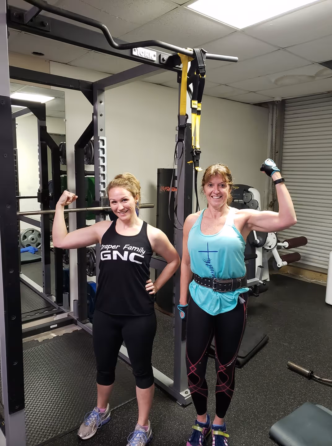 Two women flexing their arm muscles and smiling inside a gym with workout equipment.