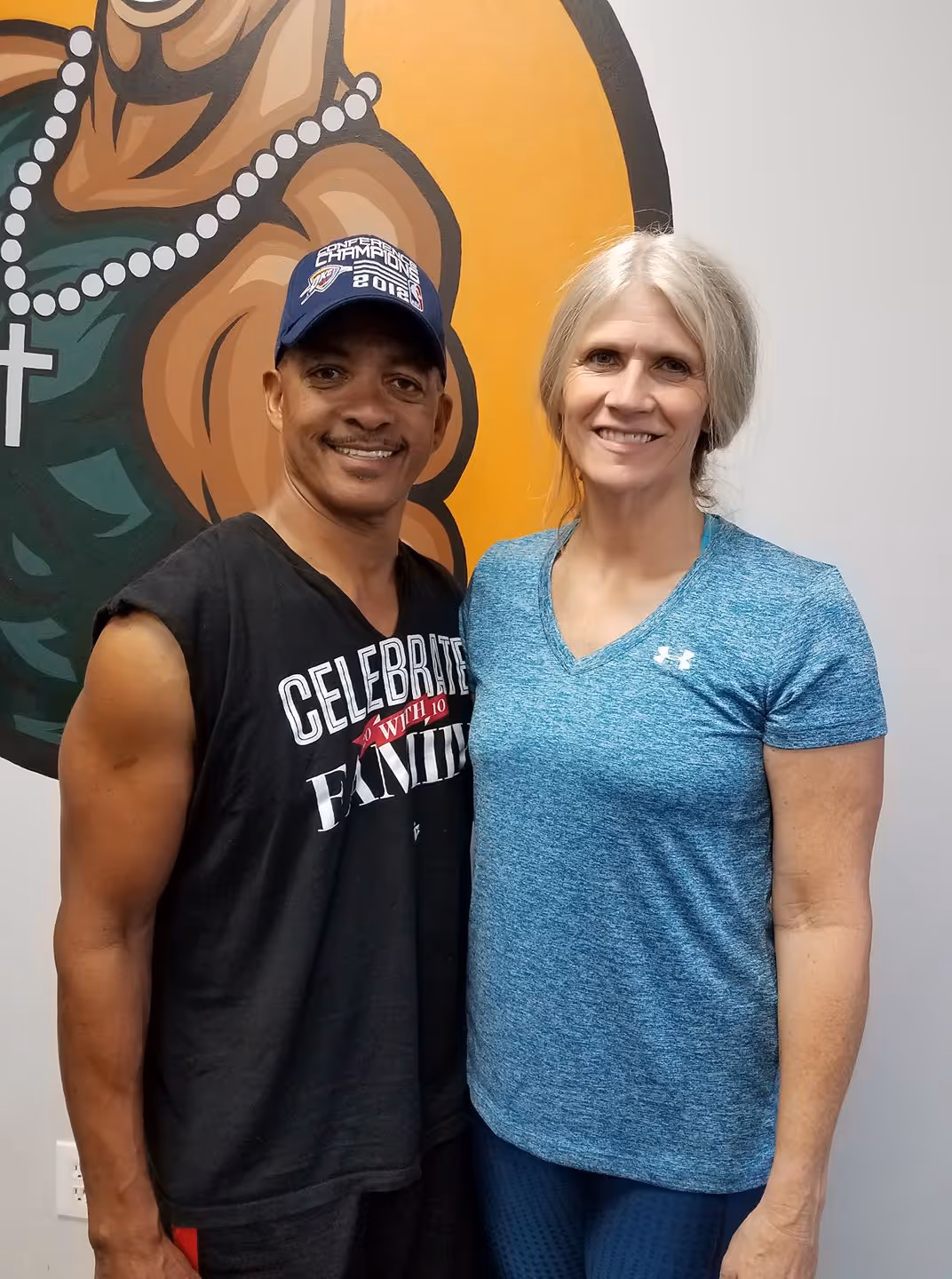 Man wearing black sleeveless shirt and cap stands next to smiling woman in blue shirt indoors.