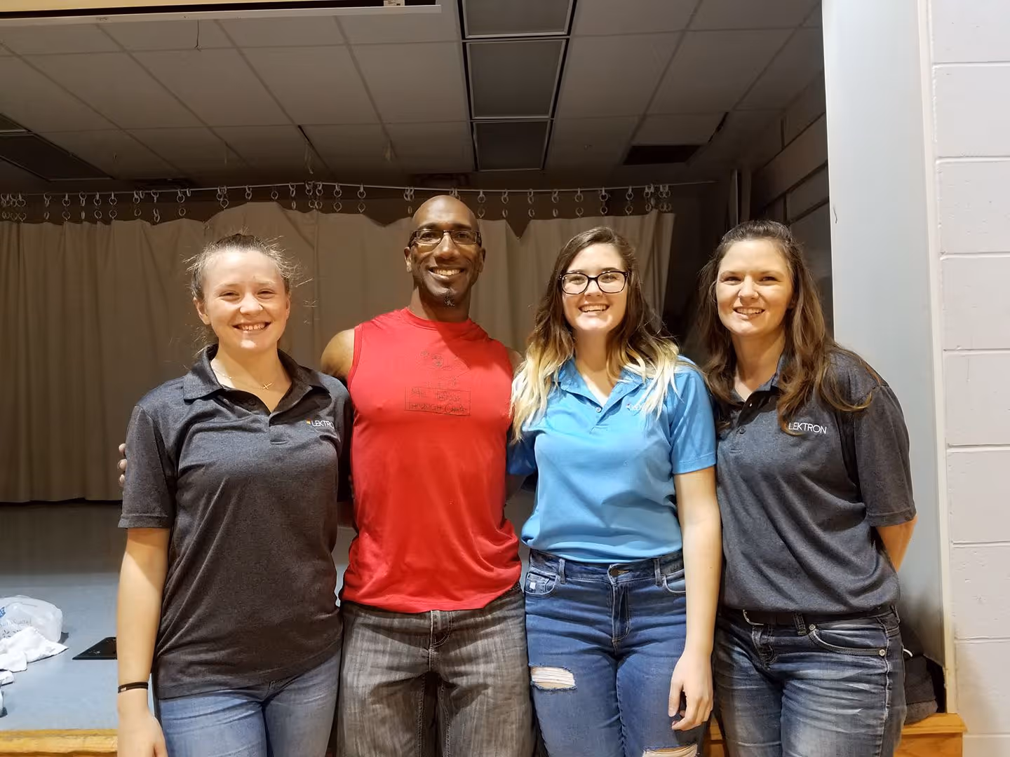 Four smiling people standing indoors, with two women wearing gray shirts and one woman in blue shirt.