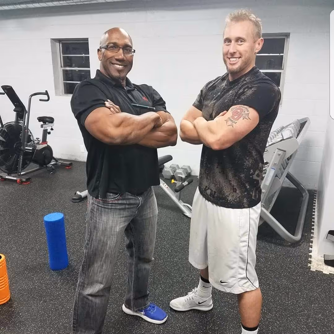 Two men with muscular arms crossed, smiling, standing in a gym with exercise equipment around.
