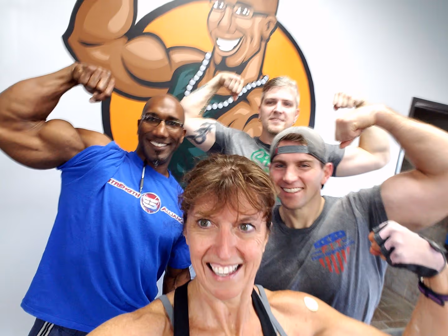 Four smiling people flexing their muscles in a gym with a cartoon muscular figure on the wall behind.