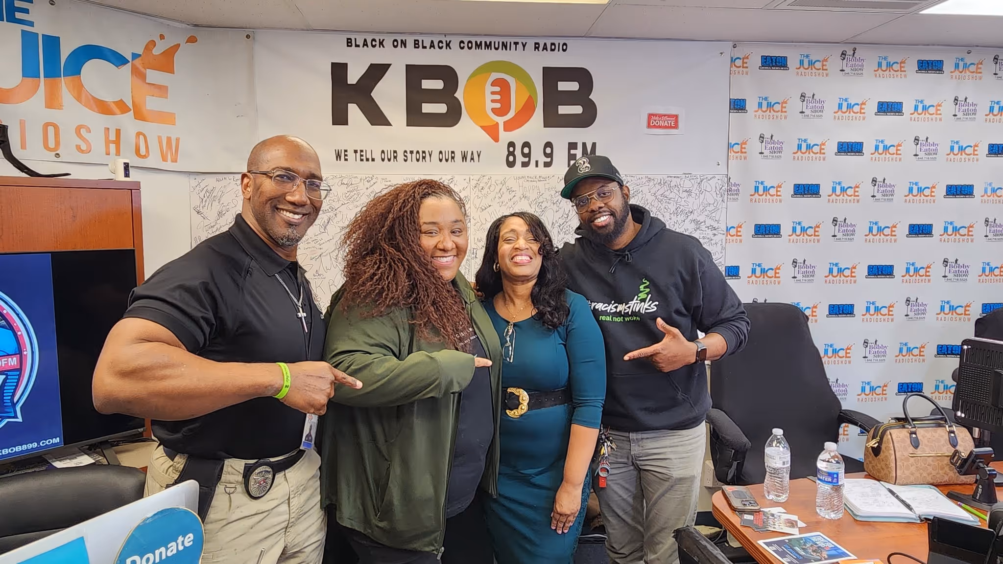 Four smiling adults standing close inside a radio station studio with KBOB 89.9 FM banner behind.