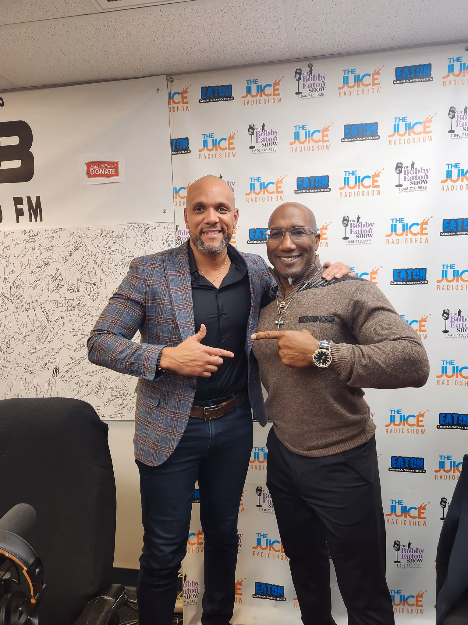 Two men smiling and pointing at each other in a radio show studio with branded backdrops.