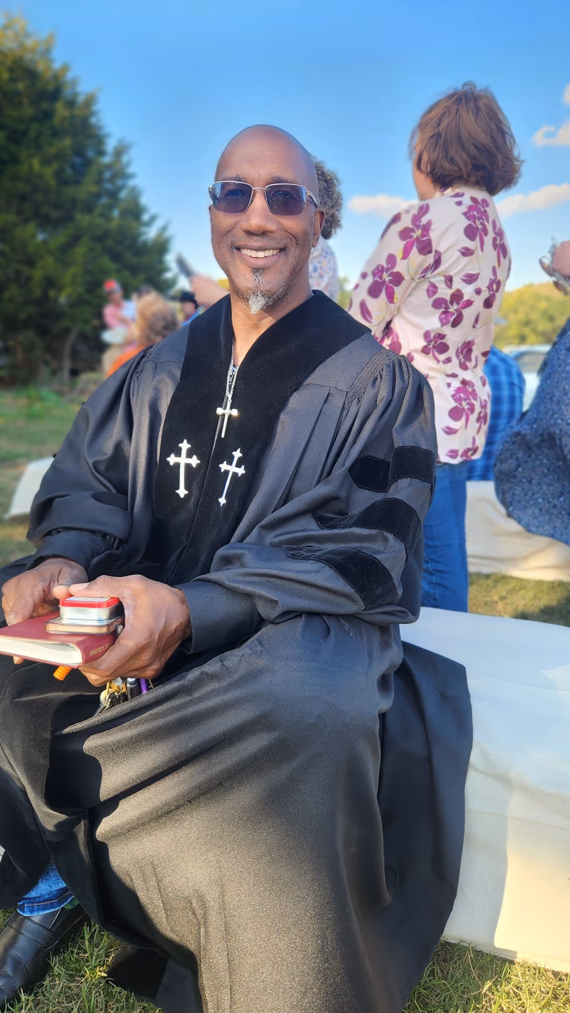 Smiling man in black clerical robe with cross symbols seated outdoors, holding books and glasses.