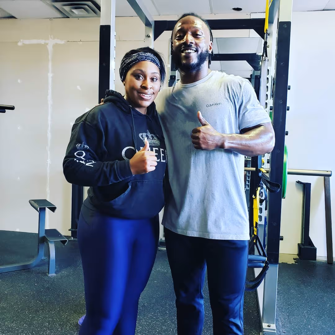 Two people in gym wear giving thumbs up and smiling, standing near weightlifting equipment.