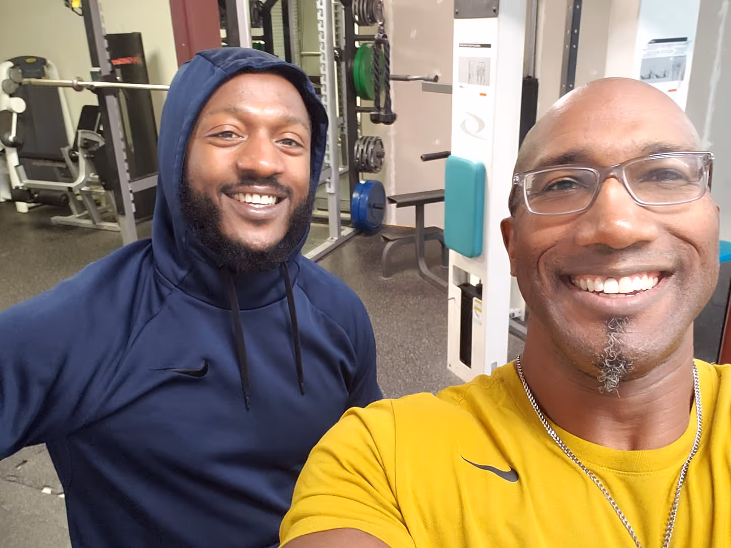 Two men smiling for a selfie in a gym with weight machines in the background.