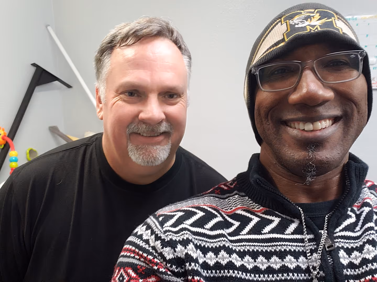Two men smiling; one wearing a patterned sweater and hat, the other in a plain black shirt.