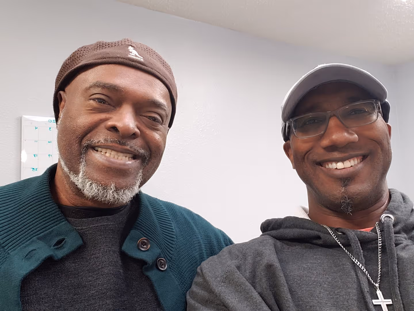 Two men smiling indoors; one wears a brown hat and green sweater, the other a gray hoodie and cap.