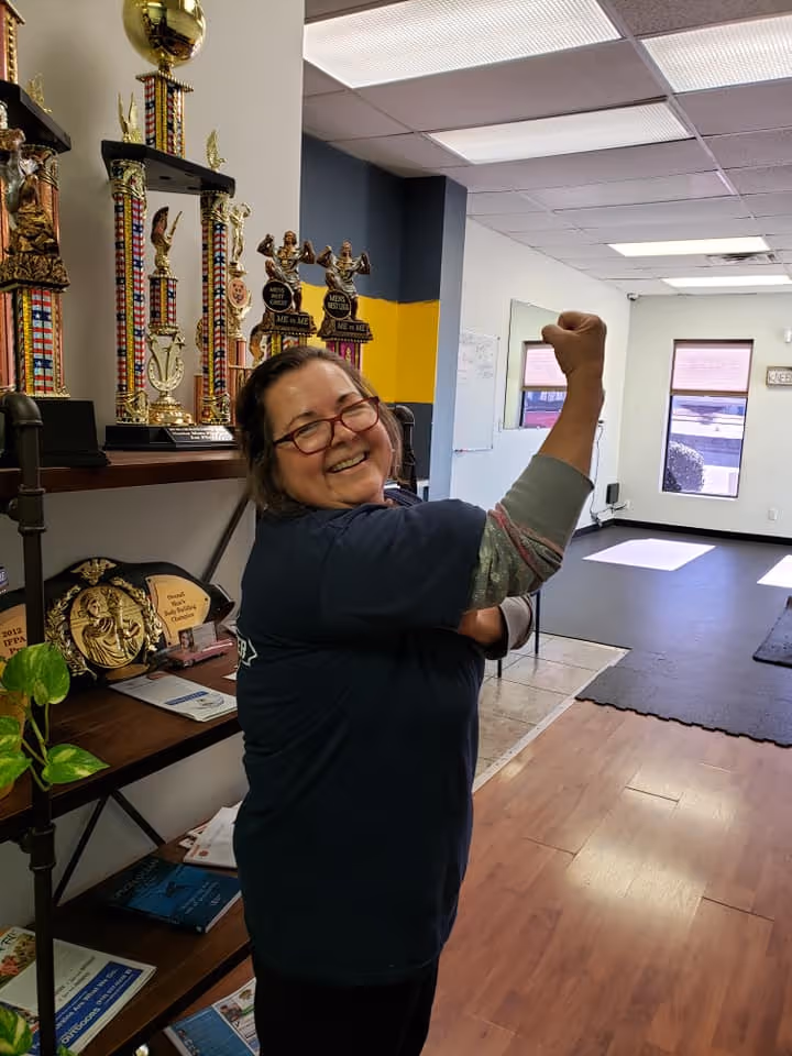 Smiling woman with glasses flexes arm in room with wooden floor and shelves of trophies.