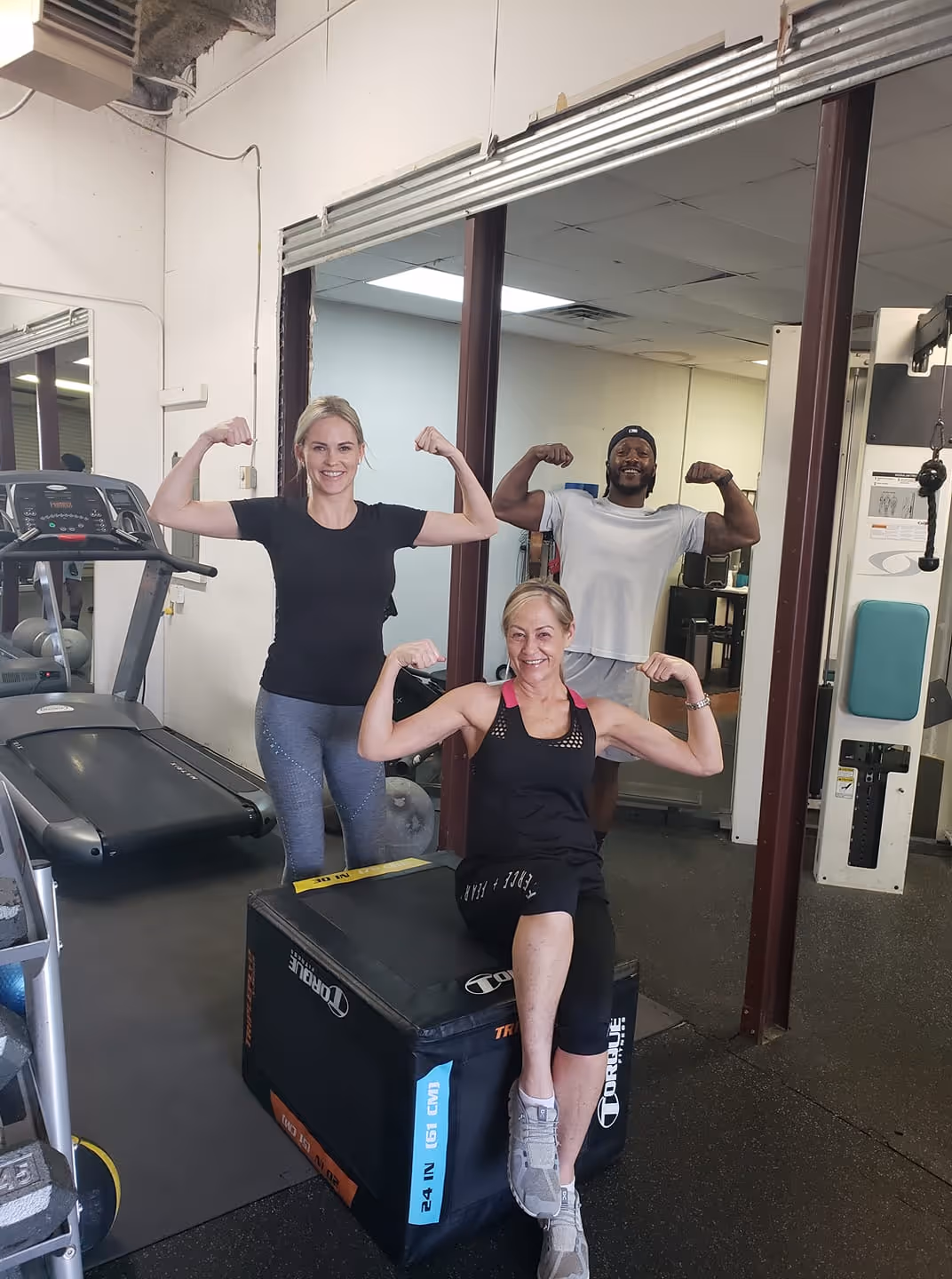 Three people in gym flexing their arm muscles and smiling near exercise equipment.