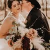 Bride holding bouquet while groom in hat kisses her cheek during outdoor wedding portrait.