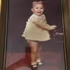 Young child standing in a white outfit with white socks and shoes against a dark background.