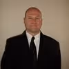 Bald man in a black suit and tie against a plain light background.