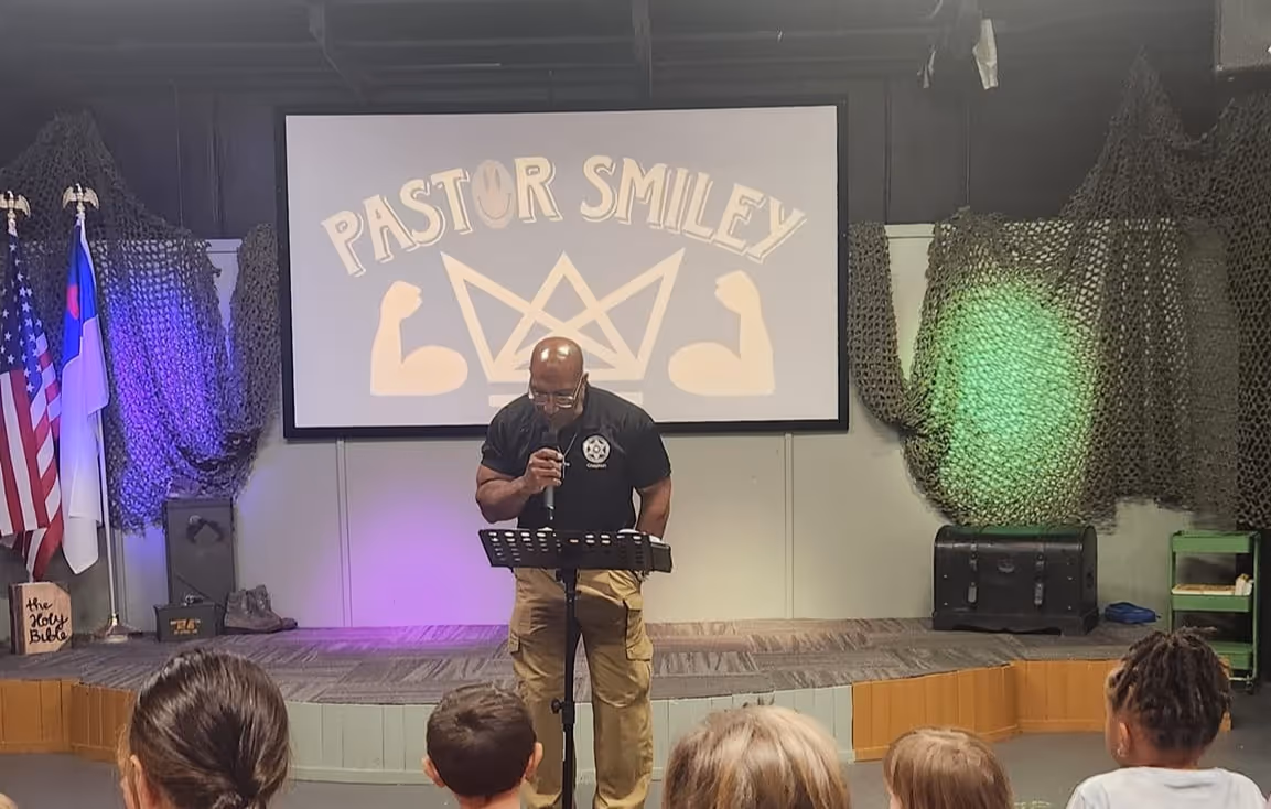 Pastor speaking to a small audience on a stage with 'Pastor Smiley' displayed on screen behind him.