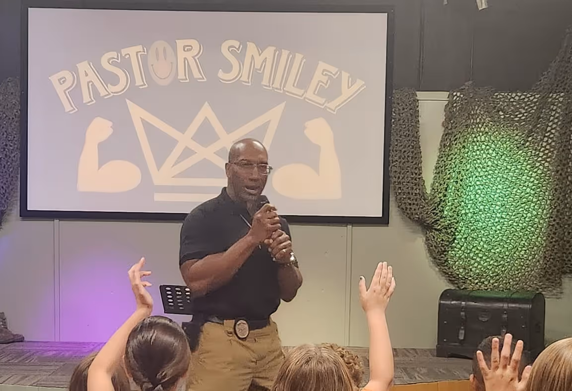 Pastor Smiley speaking to an audience with raised hands in front of a screen showing his name and logo.